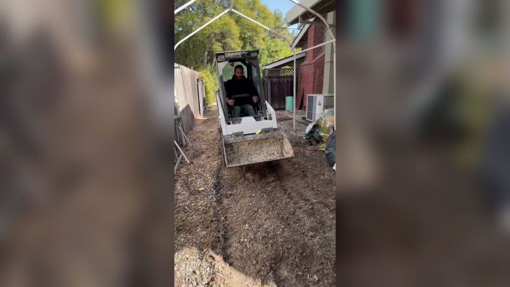 Compact Bobcat skid steer with an operator driving down a narrow side yard between a wooden fence and a house, under a temporary canopy frame