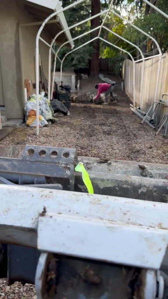 Looking down a narrow side yard from behind the bucket of a skid steer, wooden fence on the left and a canopy frame overhead