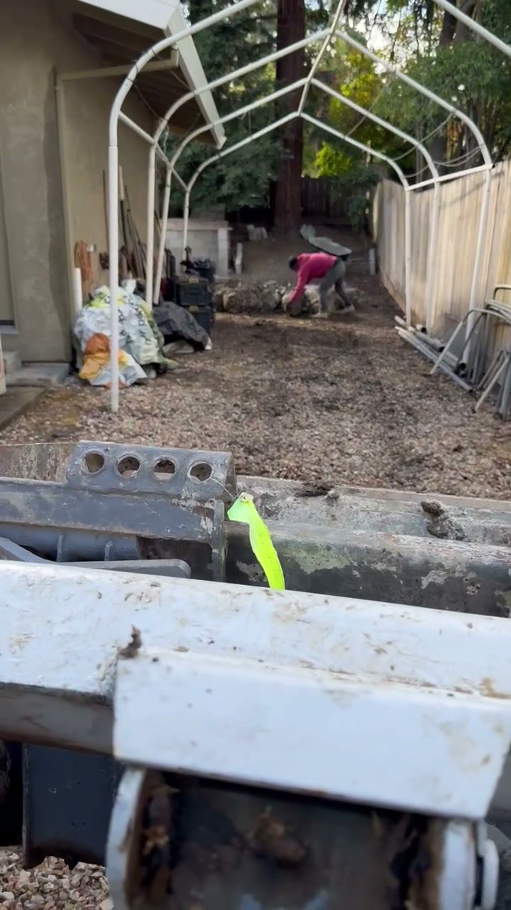 Looking down a narrow side yard from behind the bucket of a skid steer, wooden fence on the left and a canopy frame overhead