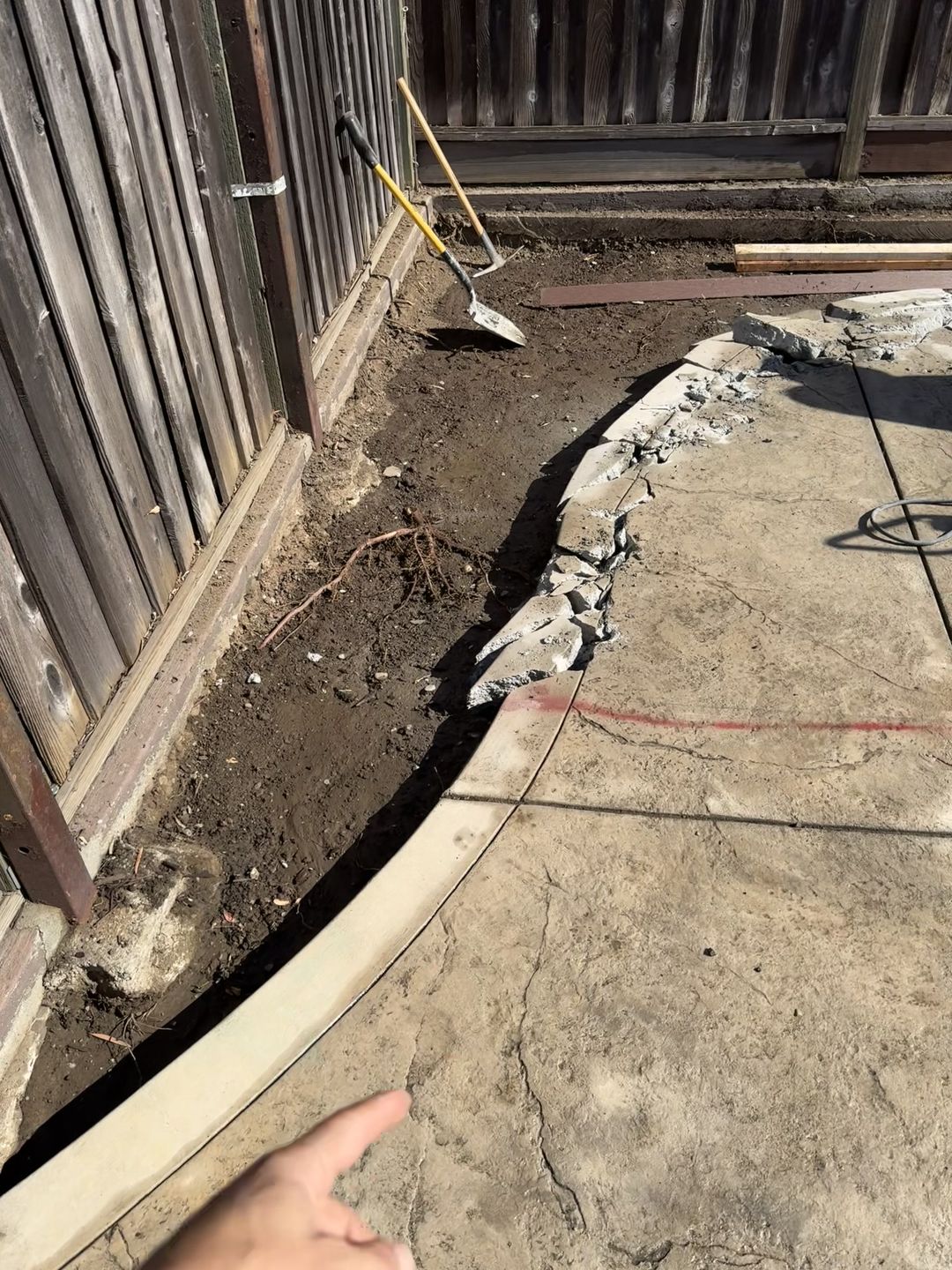Broken edge of a stamped concrete slab beside a wooden fence with exposed soil and a shovel