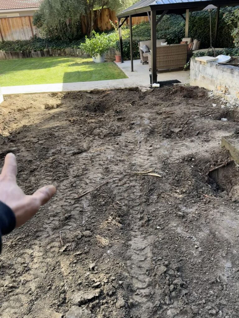 Freshly worked yard soil with machine track marks and a gazebo and lawn in the background
