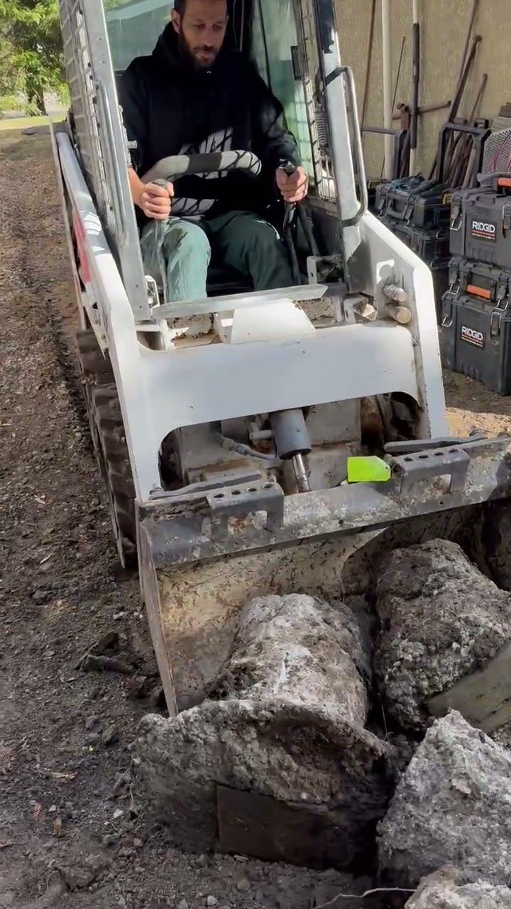 Front view of a white Bobcat skid steer with the operator seated, carrying large pieces of broken concrete in the bucket