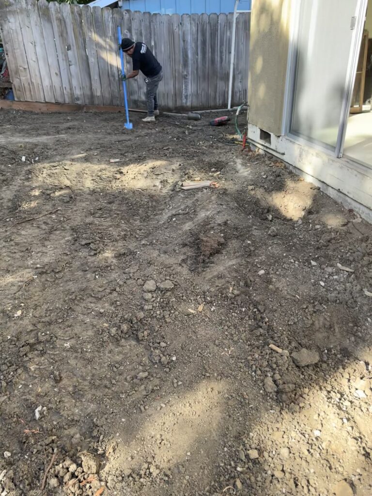Quartz crew member tamping soil level with a stucco house foundation, a crawl space vent visible just above grade, bare graded dirt sloping away from the wall